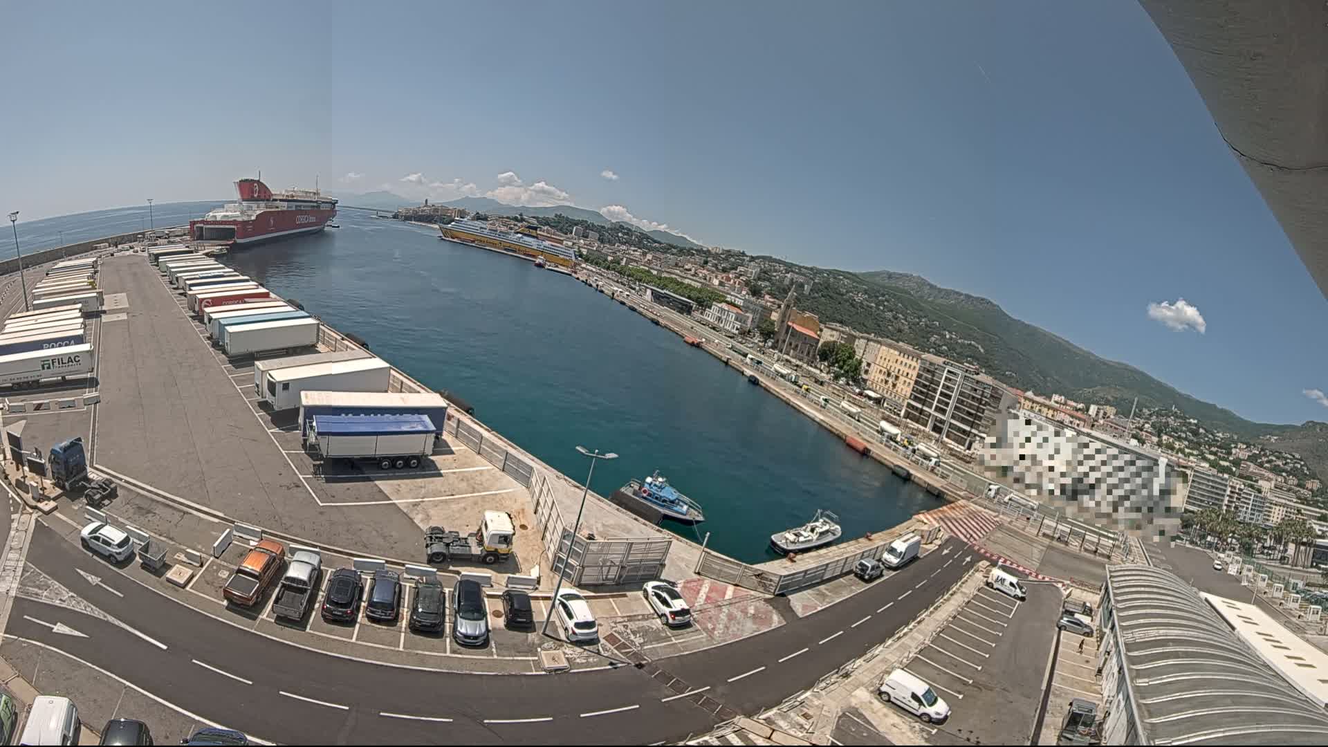 A harbor with two ferries docked, numerous parked trucks and cars, and several small boats, under a clear sunny sky.