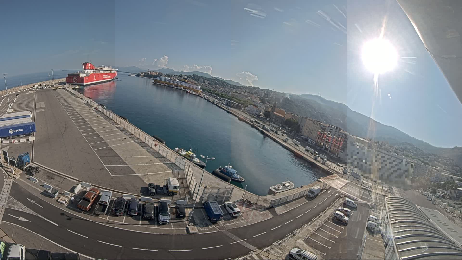 A large red ferry is docked at a port on a sunny day, with other smaller boats and vehicles visible in the harbor and parking lots surrounding the water.