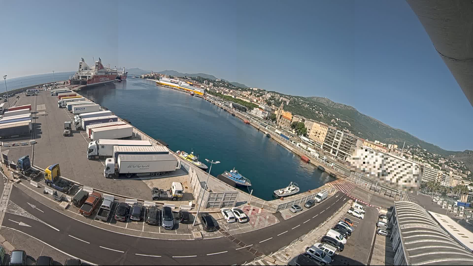 A high-angle, wide view on a sunny day shows a harbor with docked ferries, numerous trucks in a parking lot, cars, and small boats, all surrounded by buildings and hills in the distance.