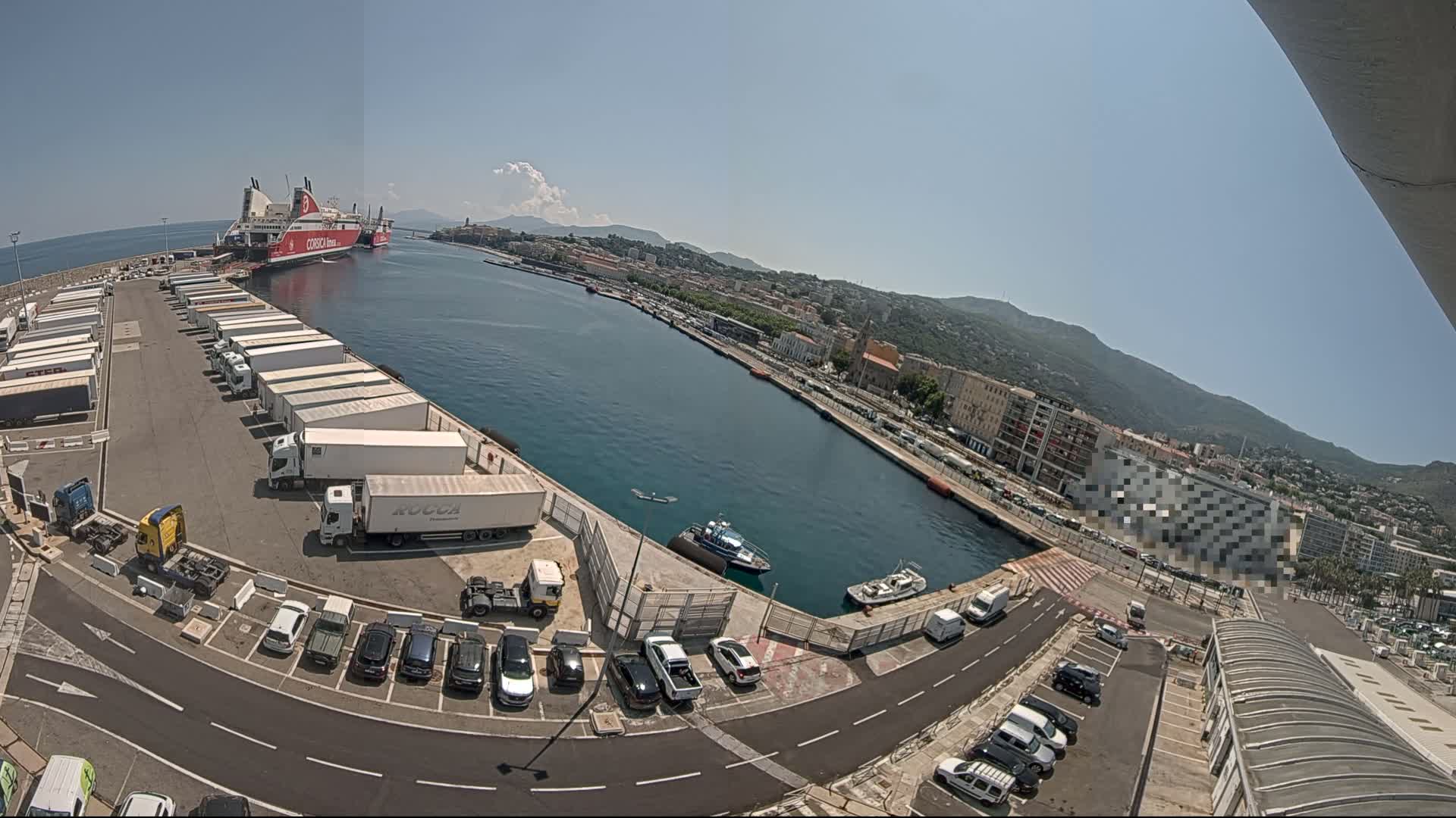 A large ferry is docked at a port city on a sunny day, with numerous trucks and cars parked nearby.