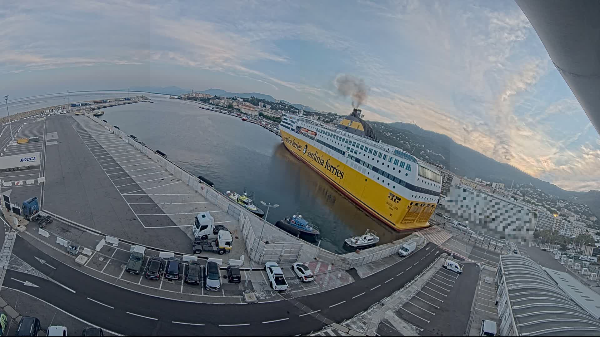 A large yellow ferry is docked at a port under a partly cloudy sky, with several vehicles parked nearby.