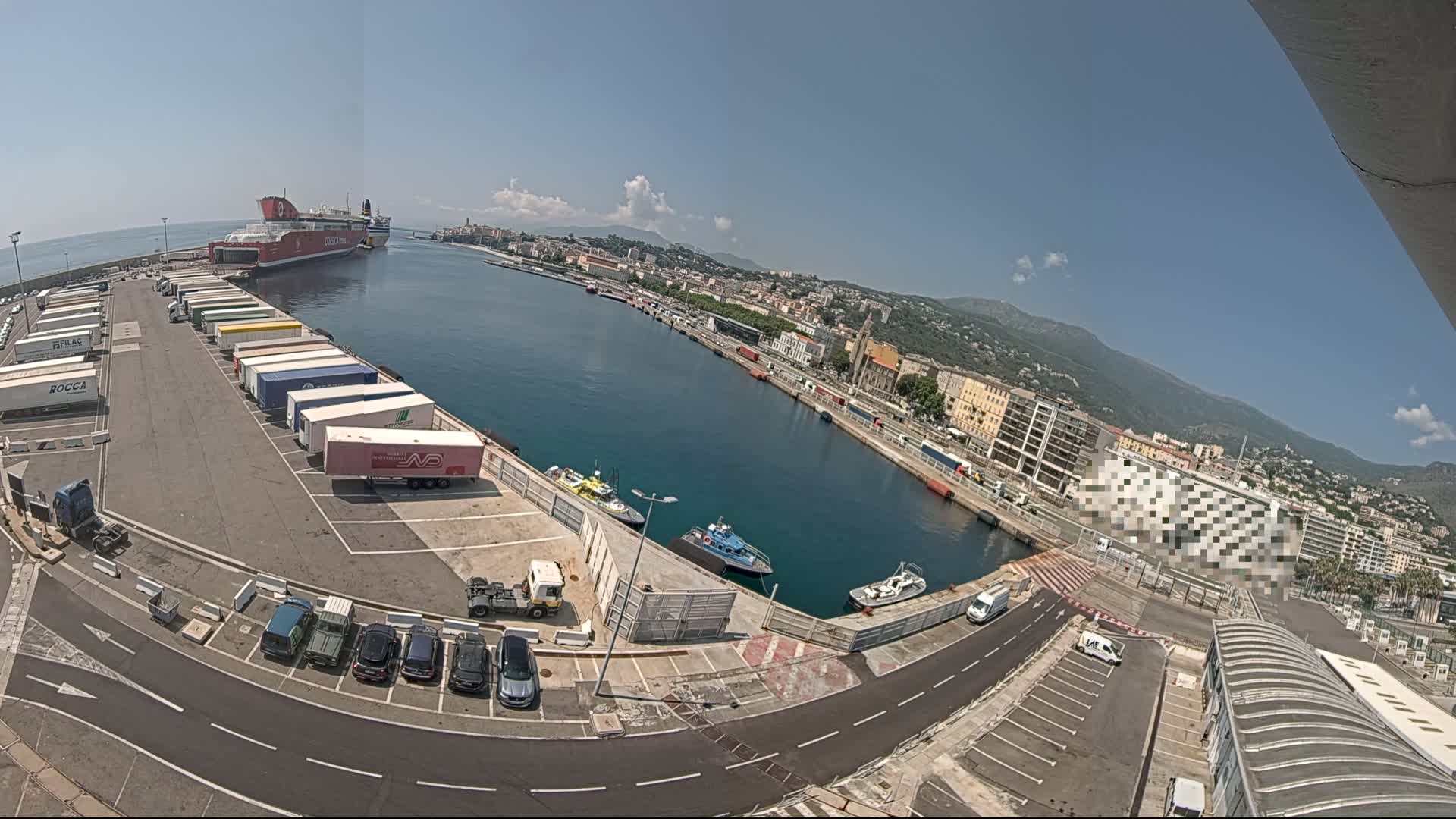A ferry is docked at a port on a sunny day, with numerous trucks and cars parked nearby, and a city and mountains visible in the background.