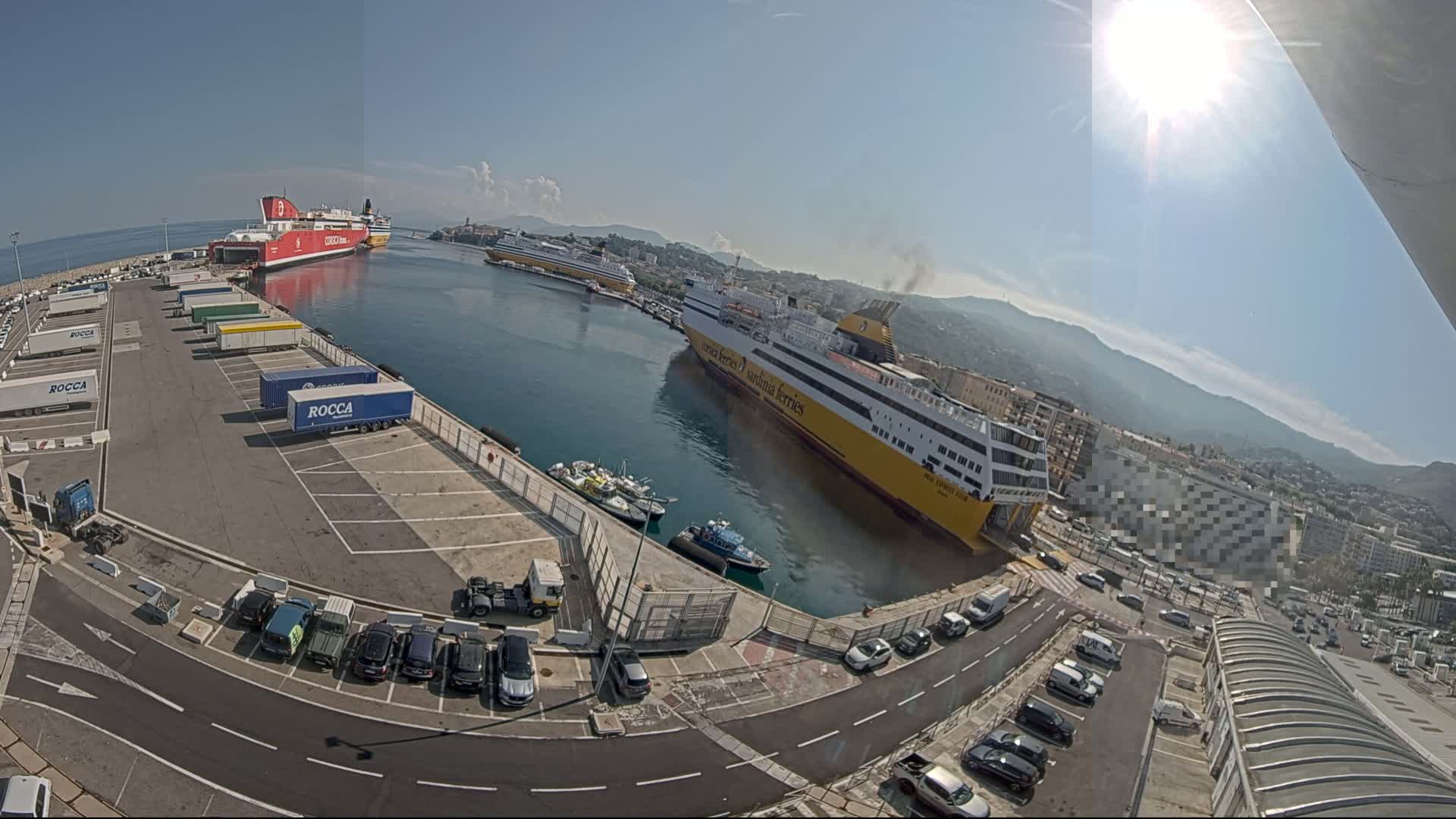 A sunny day at a harbor shows several ferries docked alongside a parking lot filled with cars and trucks.