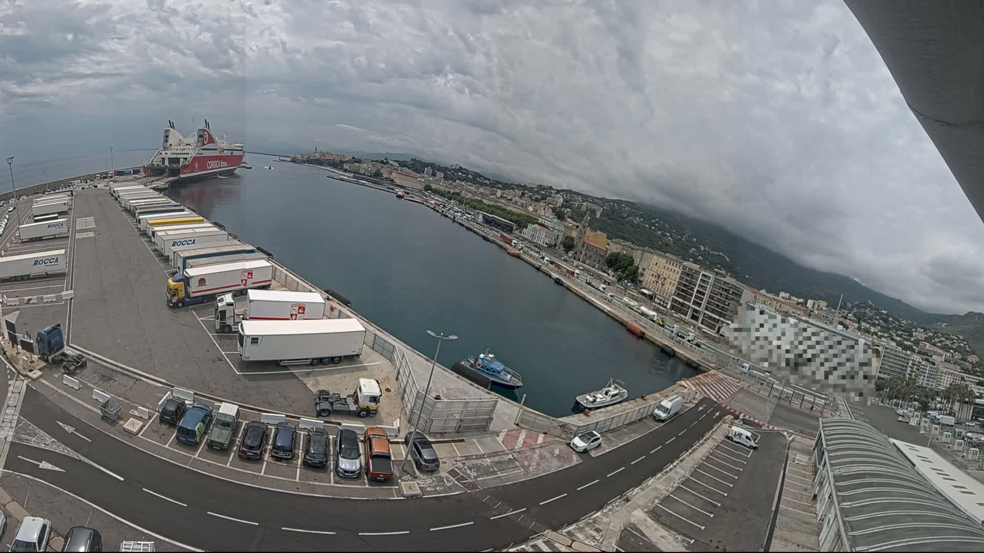 A large ferry is docked at a port under a cloudy sky, with numerous trucks and cars parked in the surrounding area.