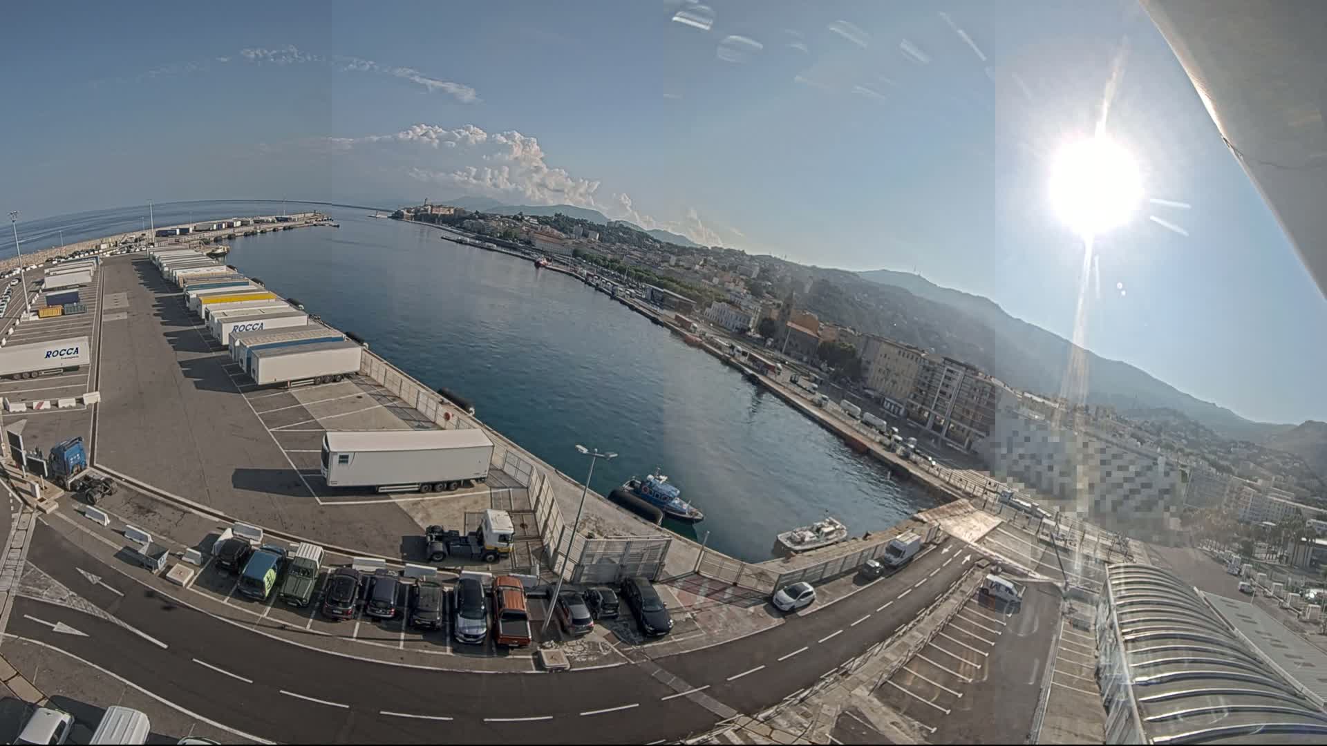 A sunny harbor scene shows a parking lot filled with trucks and cars next to a body of water with a few small boats, and a city in the background.