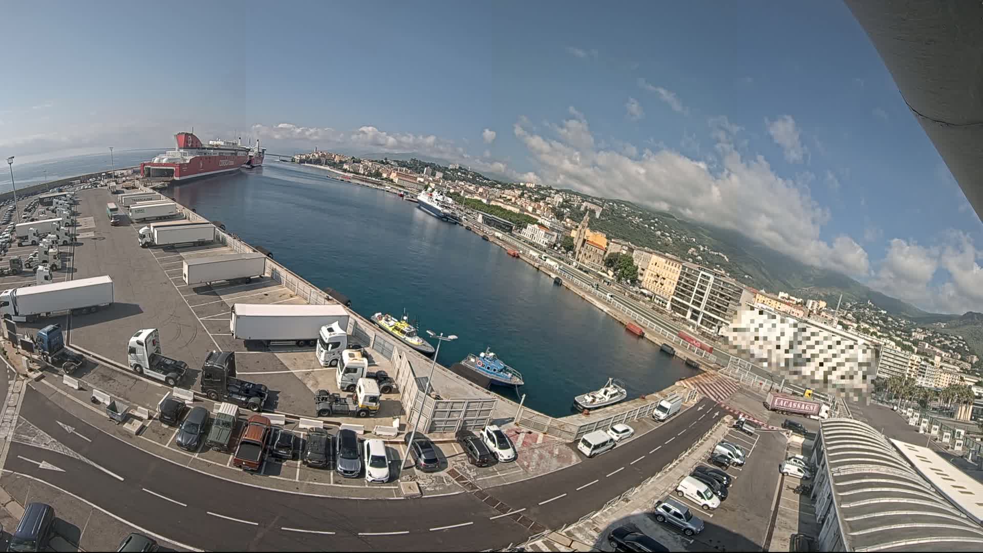 A large ferry is docked at a harbor on a partly cloudy day, with numerous trucks and cars parked in adjacent lots and smaller boats in the water.