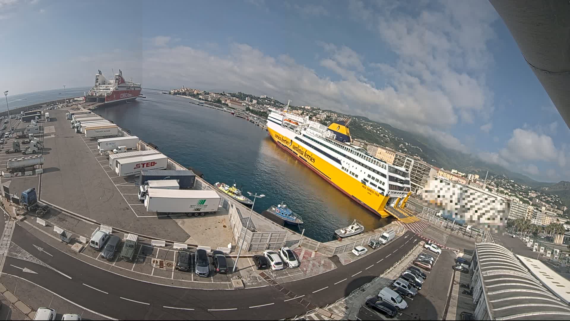 A harbor with two large ferries docked, numerous trucks and cars in a parking lot, and a city in the background under a mostly sunny sky with some clouds.