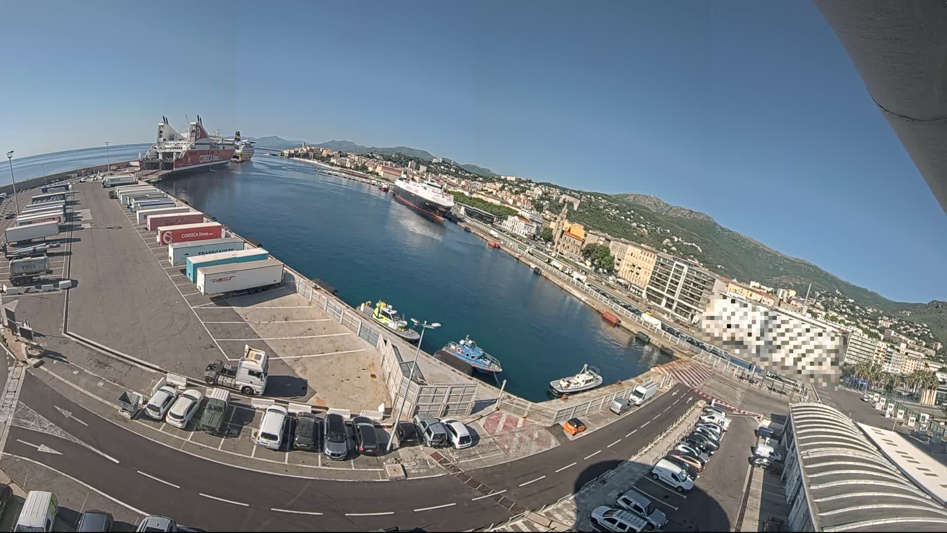 A harbor on a sunny day with a large ferry docked, numerous cargo trucks parked, smaller boats in the water, and cars in a nearby parking lot, all surrounded by buildings and hills.