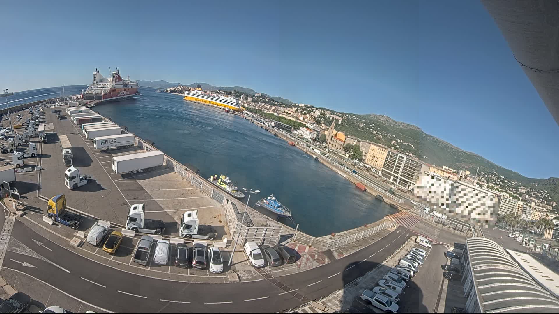 A high-angle view of a harbor on a sunny day shows two large ferries, numerous vehicles parked in a lot, and several smaller boats in the water, with a city and mountains in the background.