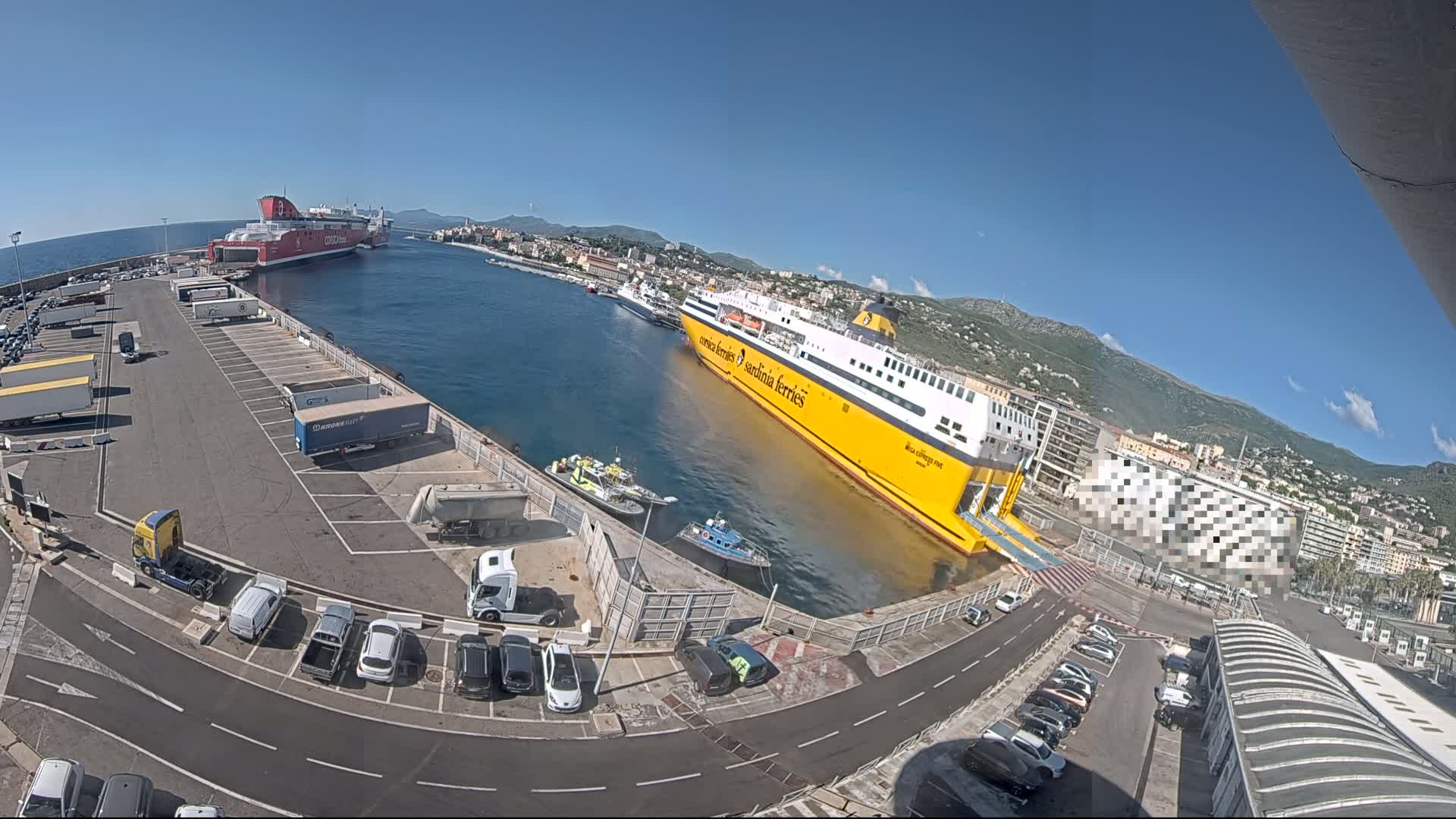 A yellow and a red ferry are docked at a port on a sunny day, with several trucks and cars parked in the surrounding lot.