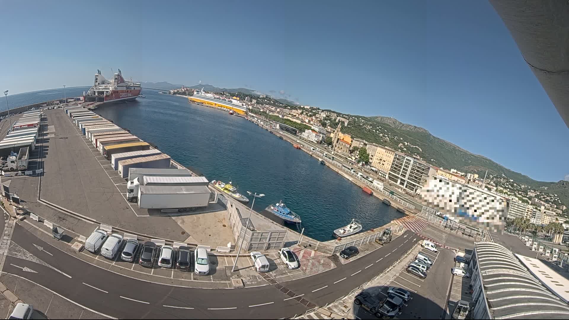 A harbor on a sunny day shows two large ferries docked, numerous trucks and cars parked along the waterfront, and smaller boats in the water.