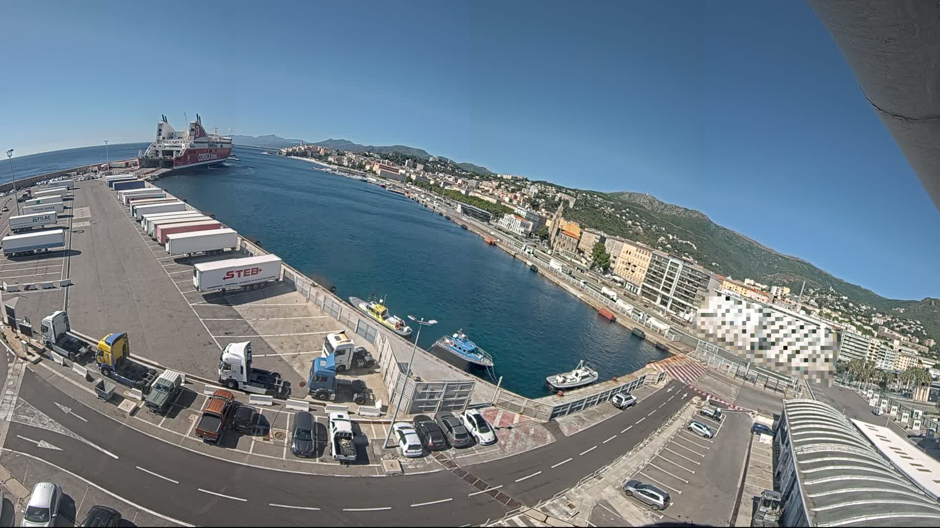 A ferry is docked at a port city on a sunny day, with numerous trucks and cars parked nearby, and several small boats in the water.
