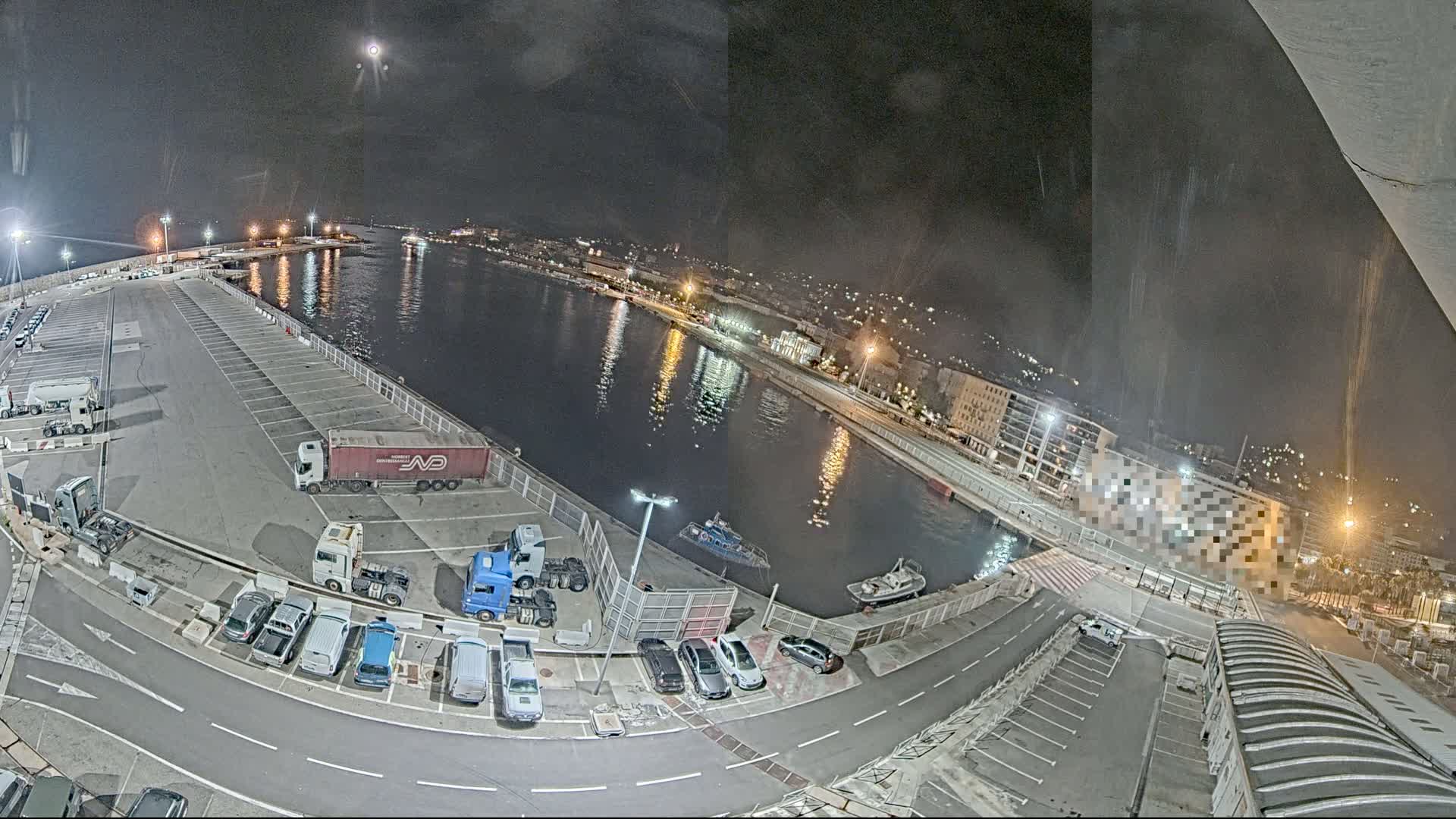 A nighttime, high-angle view shows a harbor with parked cars and trucks, a calm body of water reflecting lights, and a city skyline in the background under a clear, moonlit sky.