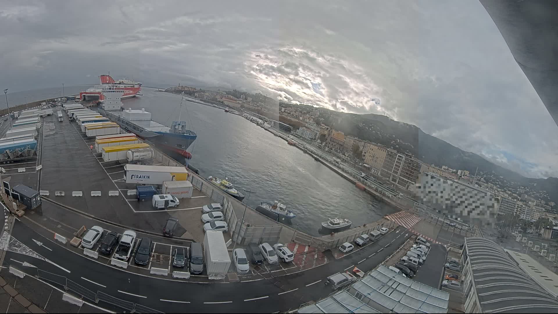 A nighttime, high-angle view shows a harbor with parked cars and trucks, a calm body of water reflecting lights, and a city skyline in the background under a clear, moonlit sky.