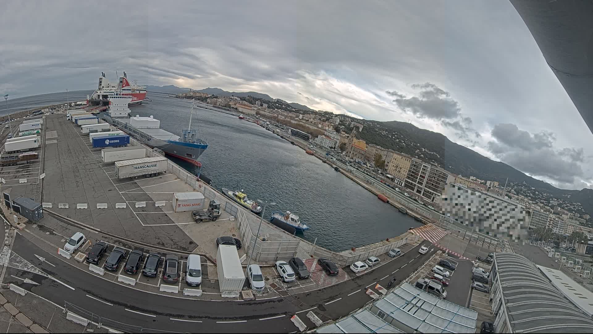 A wide-angle view captures a bustling port on an overcast day, featuring several large ships docked alongside piers filled with truck trailers and parked cars, with a city and green mountains lining the distant shore under a cloudy sky.