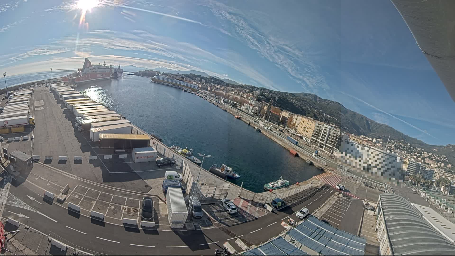 A wide-angle view captures a bustling port on an overcast day, featuring several large ships docked alongside piers filled with truck trailers and parked cars, with a city and green mountains lining the distant shore under a cloudy sky.