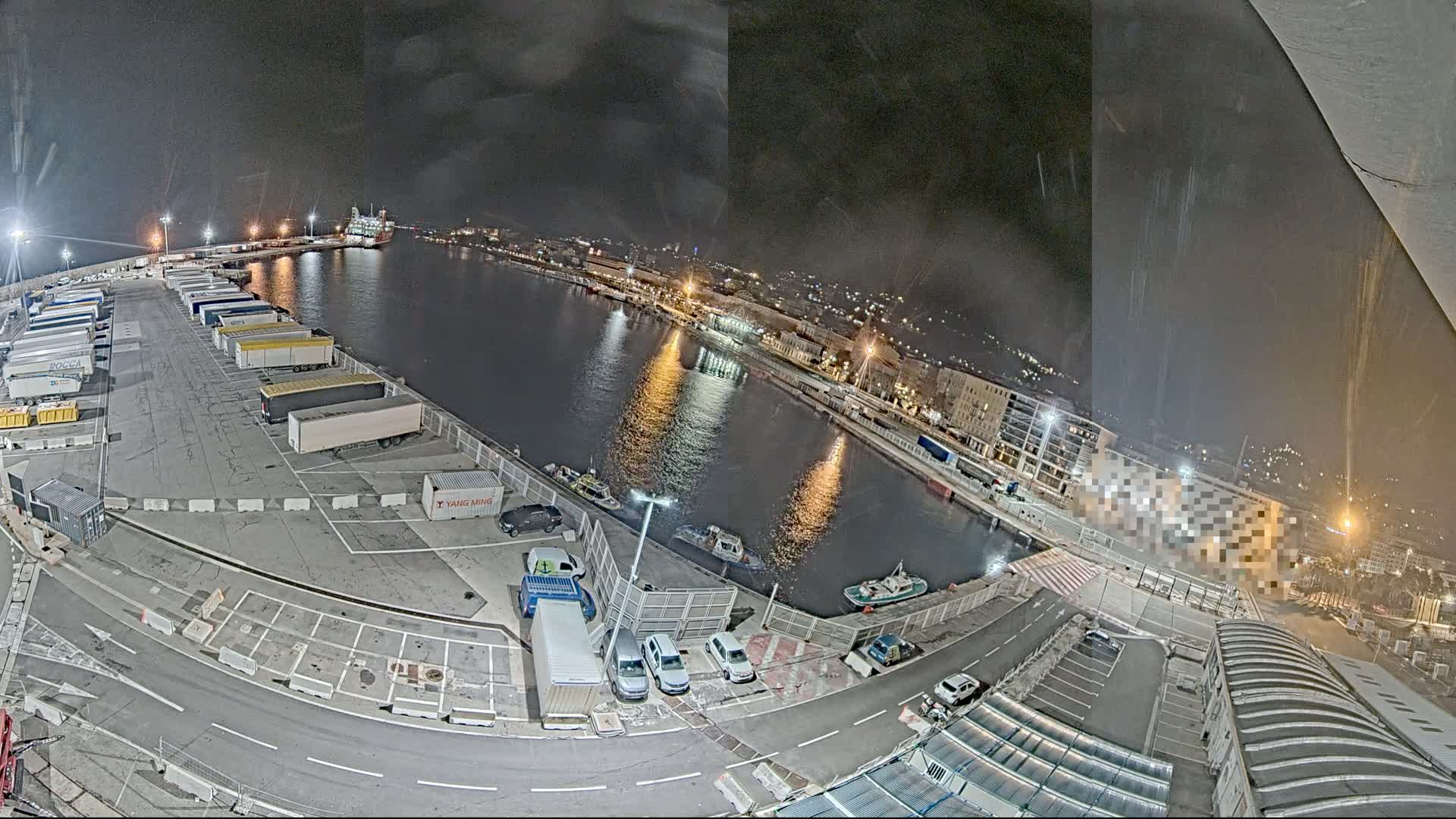 A wide-angle view captures a bustling port on an overcast day, featuring several large ships docked alongside piers filled with truck trailers and parked cars, with a city and green mountains lining the distant shore under a cloudy sky.