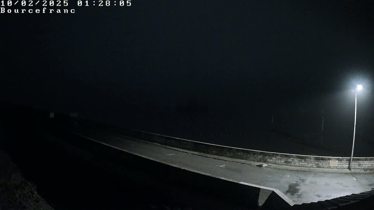A dark, foggy night scene shows a roadway curving alongside a low wall, illuminated by a single streetlight.