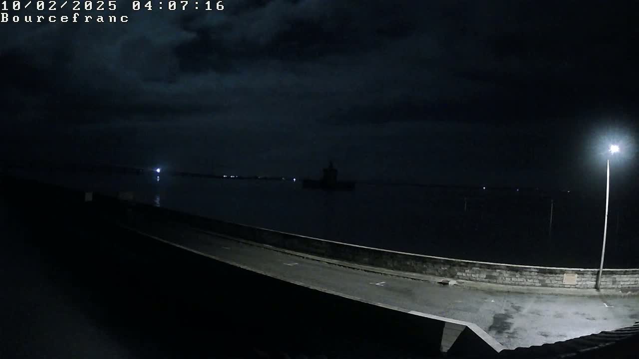 A nighttime view of a dark body of water under a cloudy sky, with a paved area and a single lit lamp in the foreground.