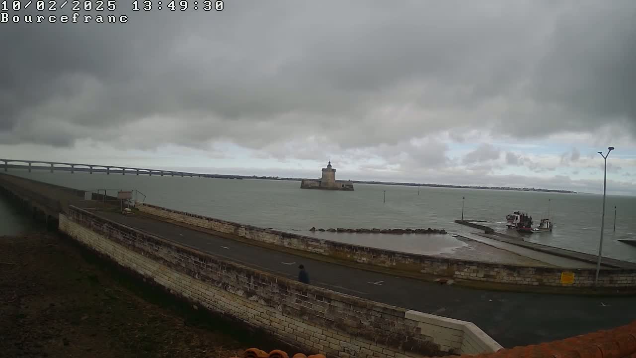 A stone seawall with a road curves around a small harbor where a boat is docked, overlooking a calm body of water with a distant bridge and a stone tower under an overcast sky.