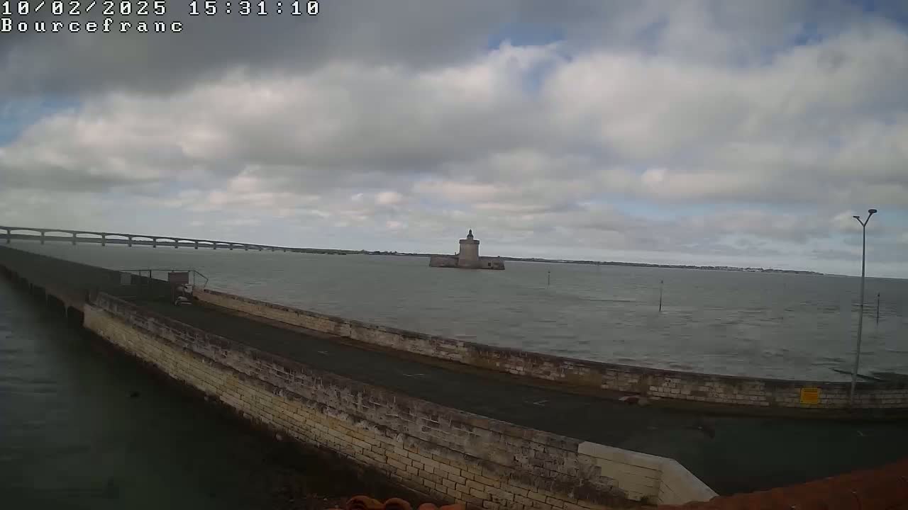 A stone causeway extends into a choppy sea toward a small, stone tower, with a long bridge visible in the distance under a cloudy sky.