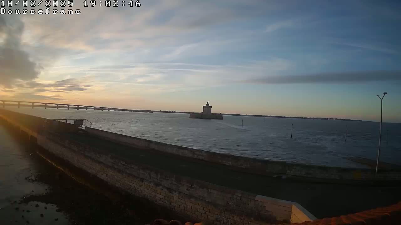 A stone wall borders a calm body of water at sunset, with a distant bridge and a small stone tower visible in the background.