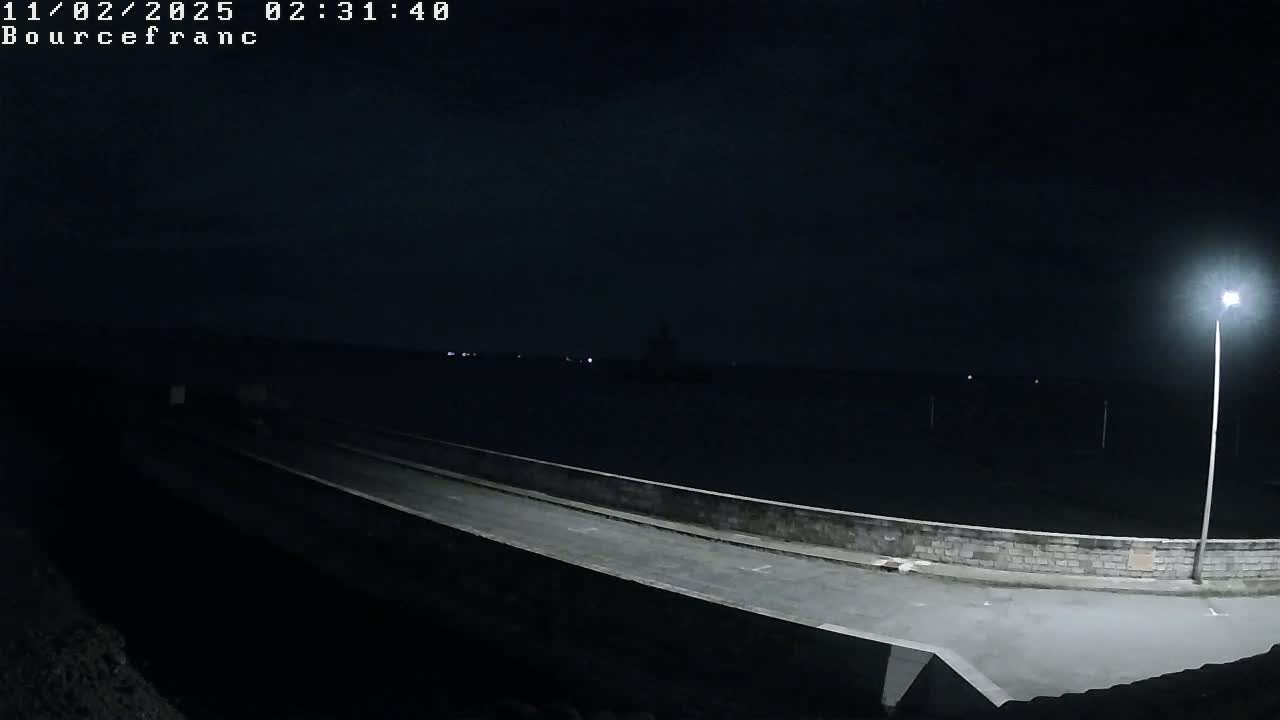 A nighttime view shows a paved pathway extending from the bottom to the middle of the image, bordered by a low stone wall, with a bright streetlight illuminating the scene and a dark body of water visible in the background under a clear night sky.