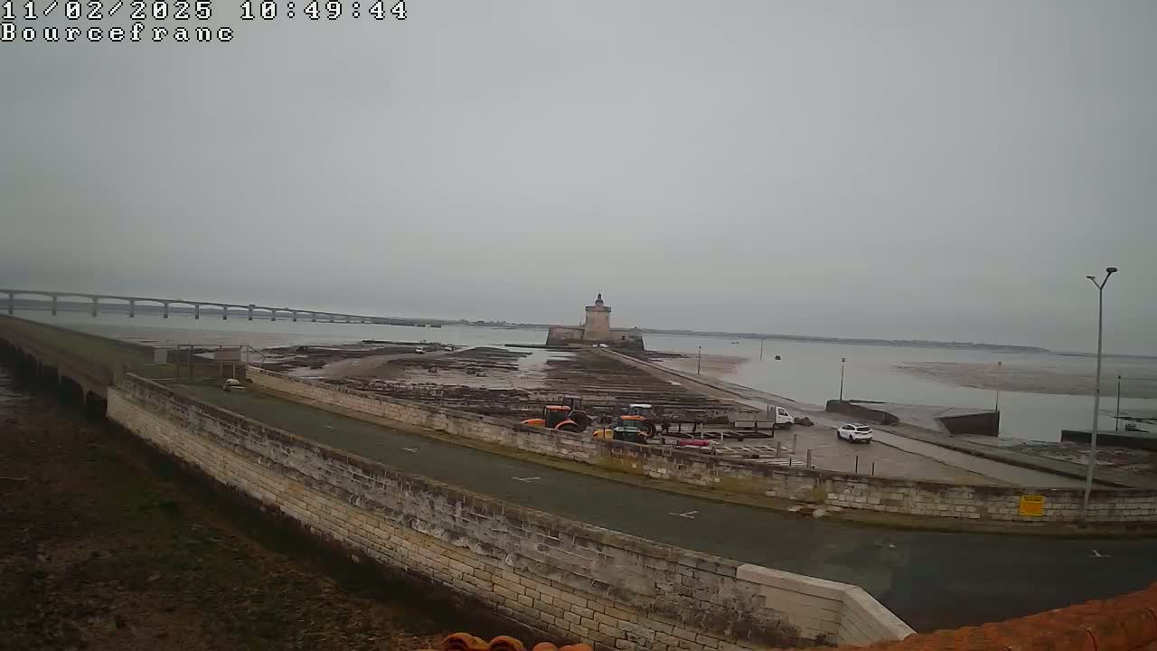 A stone wall borders a paved area with several vehicles near a tower on a tidal flat, with a long bridge visible in the distance under an overcast sky.