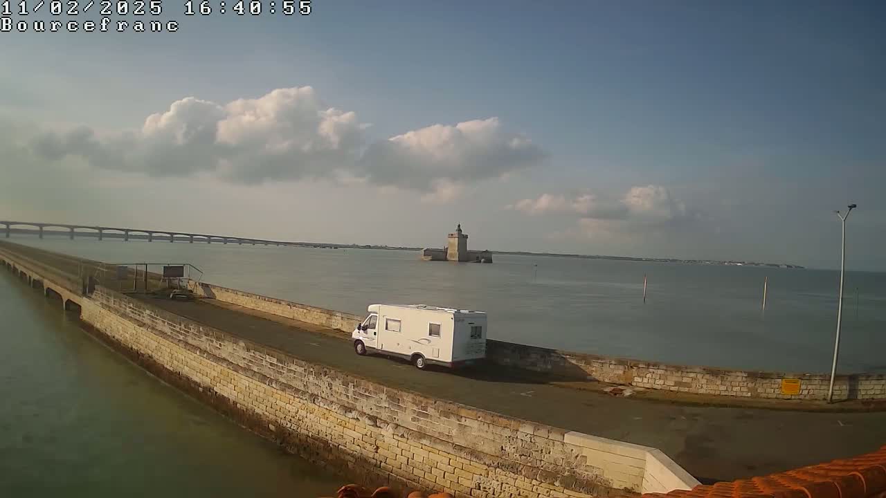 A white camper van is parked on a stone wall overlooking a calm body of water with a distant bridge and a stone tower under a partly cloudy sky.