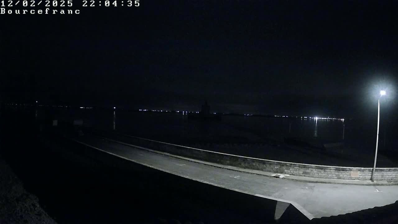 A nighttime view of a paved area next to a body of water with a distant town reflecting lights on the water under a clear, dark sky.