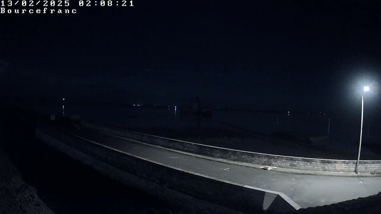 A nighttime, low-angle view shows a dark road next to a low stone wall overlooking a calm body of water under a clear, dark sky illuminated by a single lamppost.