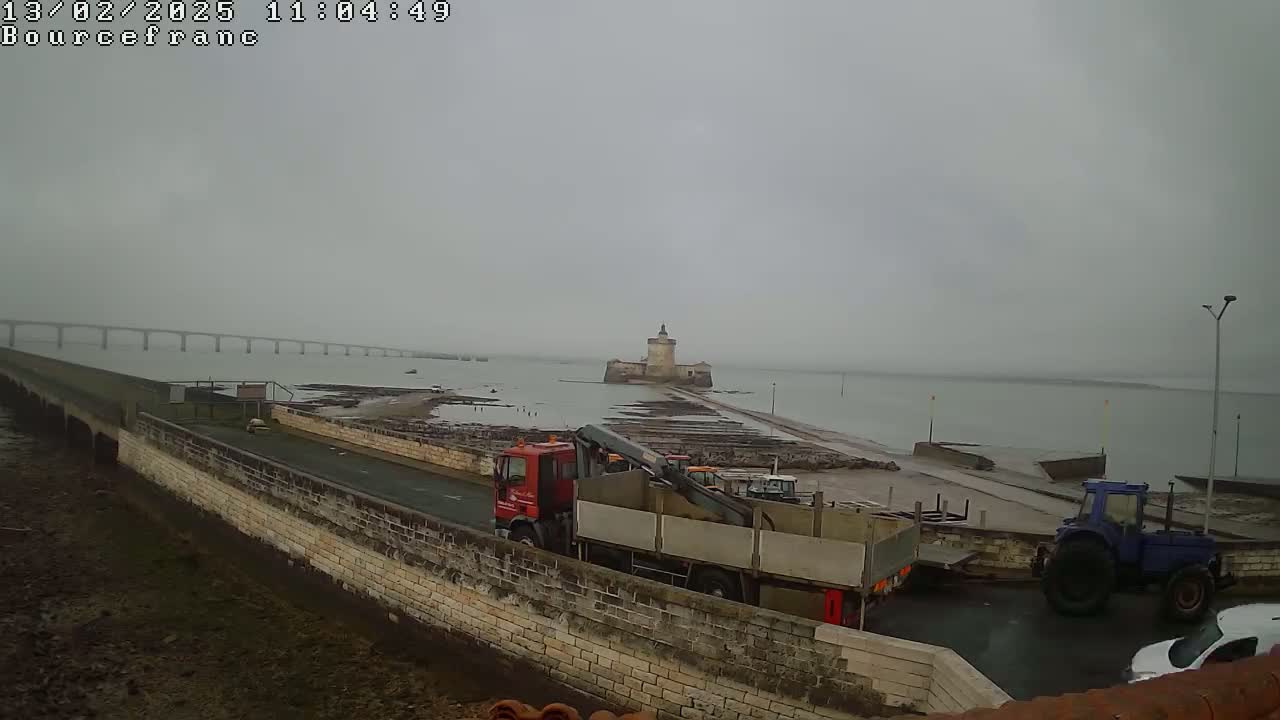 A distant bridge and a stone tower are visible across a body of water on an overcast day, with a red truck and a blue tractor near the foreground.