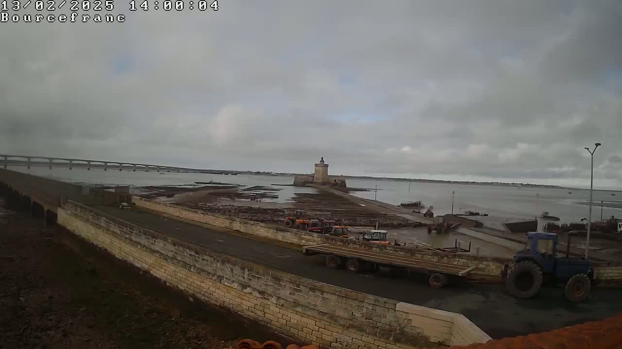A stone wall and road overlook a tidal flat with tractors and a trailer, a distant bridge, and a tower in the overcast sky.