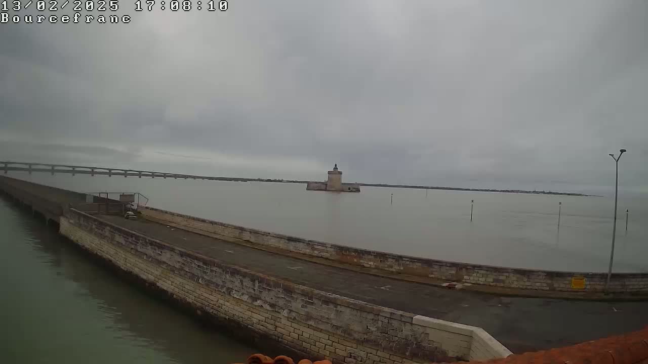 A stone wall curves around a calm body of water, under an overcast sky, with a distant bridge and a small stone tower visible in the middle ground.