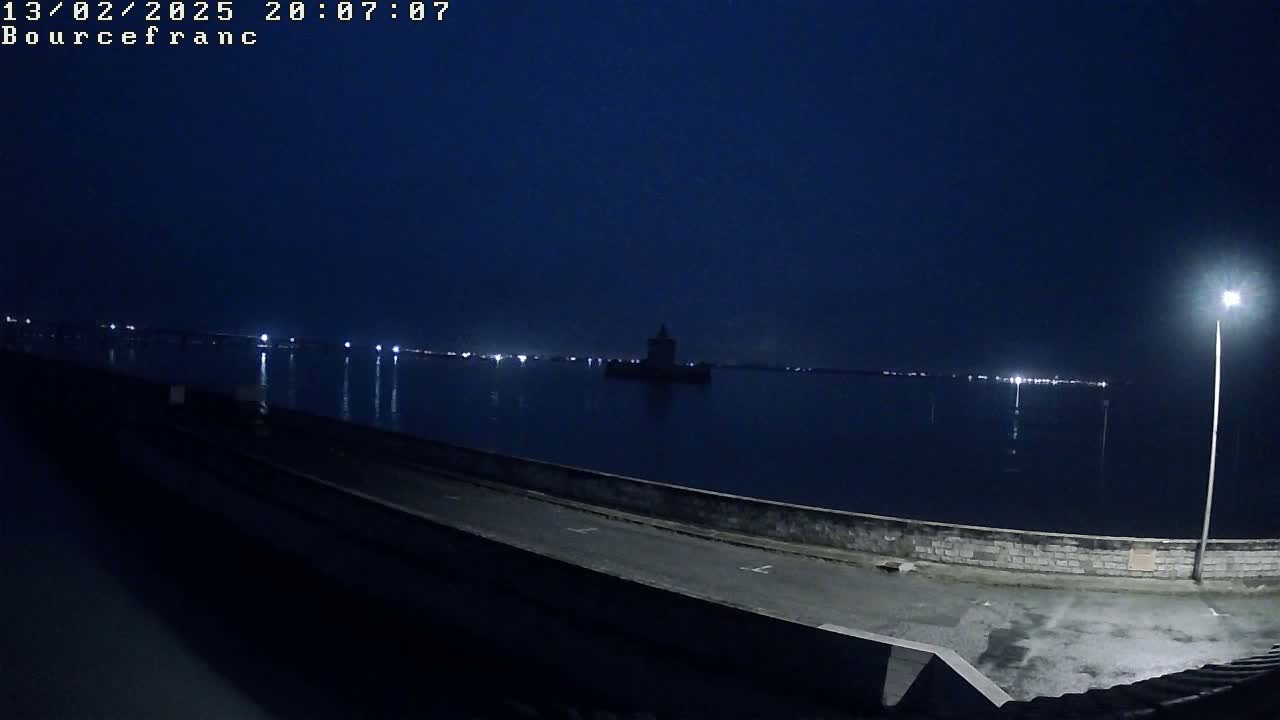 A dark, nighttime view of a calm body of water with a distant tower and a shoreline featuring a low wall and a paved road, lit by a single lamppost.