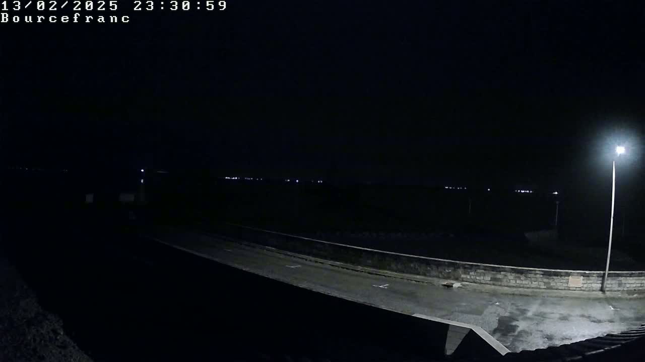 A nighttime, dimly lit outdoor scene shows a paved area next to a low wall, with a distant horizon line visible and a single bright lamp illuminating the foreground.