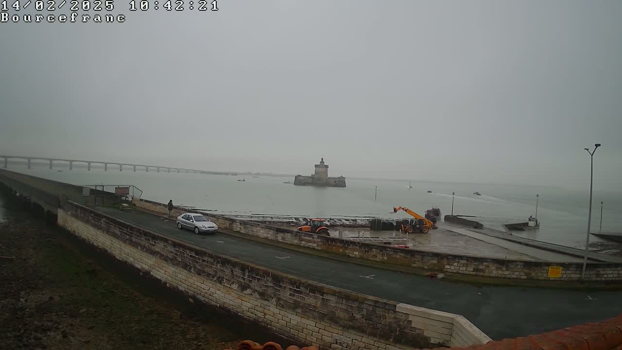 A gray, overcast day shows a stone wall and roadway next to a body of water, with a distant bridge, a small island fortress, construction equipment, and a few vehicles.