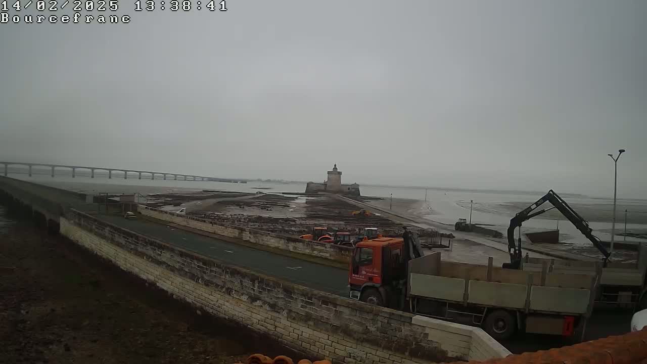 A long bridge stretches across a body of water under an overcast sky, with a stone tower visible in the middle distance, and construction vehicles near a stone wall next to a road.