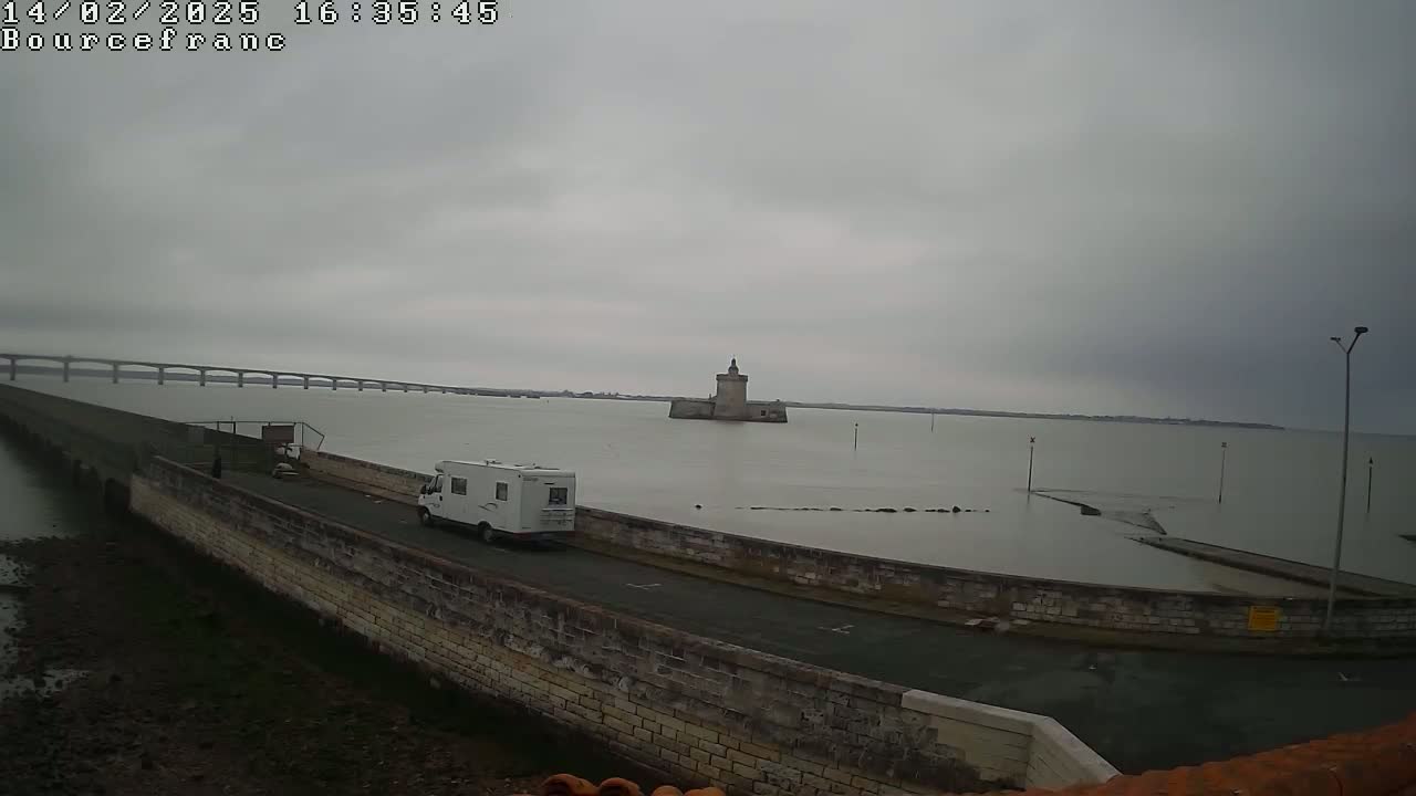A camper van is parked on a road next to a stone wall overlooking a calm body of water under an overcast sky, with a distant bridge and a stone tower visible in the background.