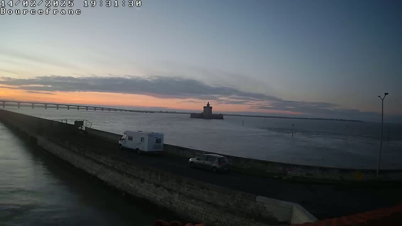 A long, low stone wall extends into a calm body of water at sunset, with a distant bridge, a stone tower, a campervan, and a car visible.