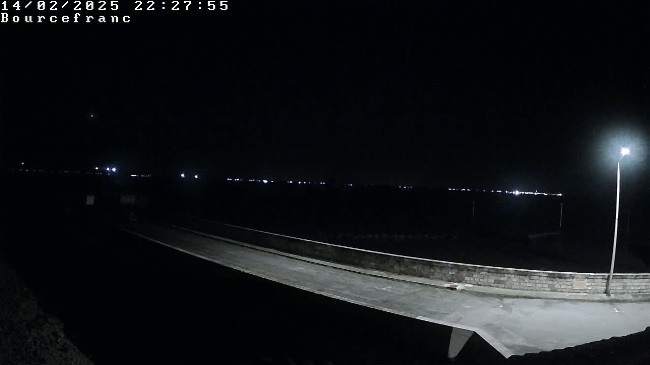 A nighttime view of a paved roadway and low wall extending into the distance, illuminated by a single lamppost and distant lights.