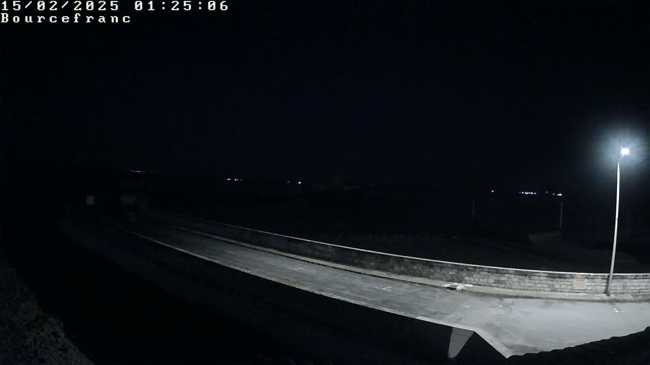 A nighttime view of an empty road on a low wall, illuminated by a single streetlight, overlooking a dark body of water.