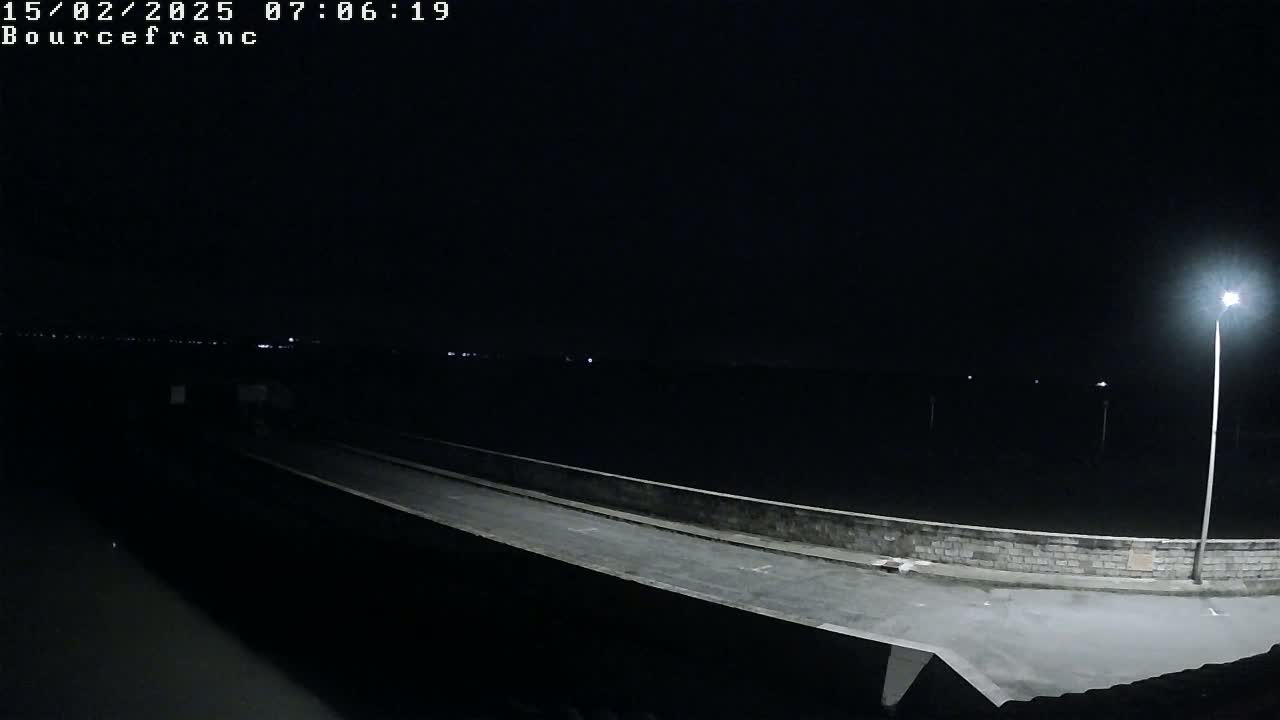 A dark, nighttime scene shows a roadway on a low wall extending into the distance under a single bright light, with a faint horizon line visible in the background.