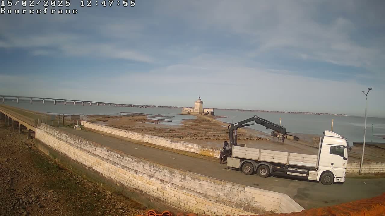 A white truck with a crane is parked on a stone road near a body of water, a stone tower is visible in the distance, and a long bridge spans the water under a partly cloudy sky.