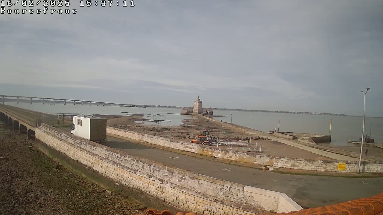 A white van drives toward a stone fort connected to the mainland by a causeway, with a bridge visible in the distance under an overcast sky.