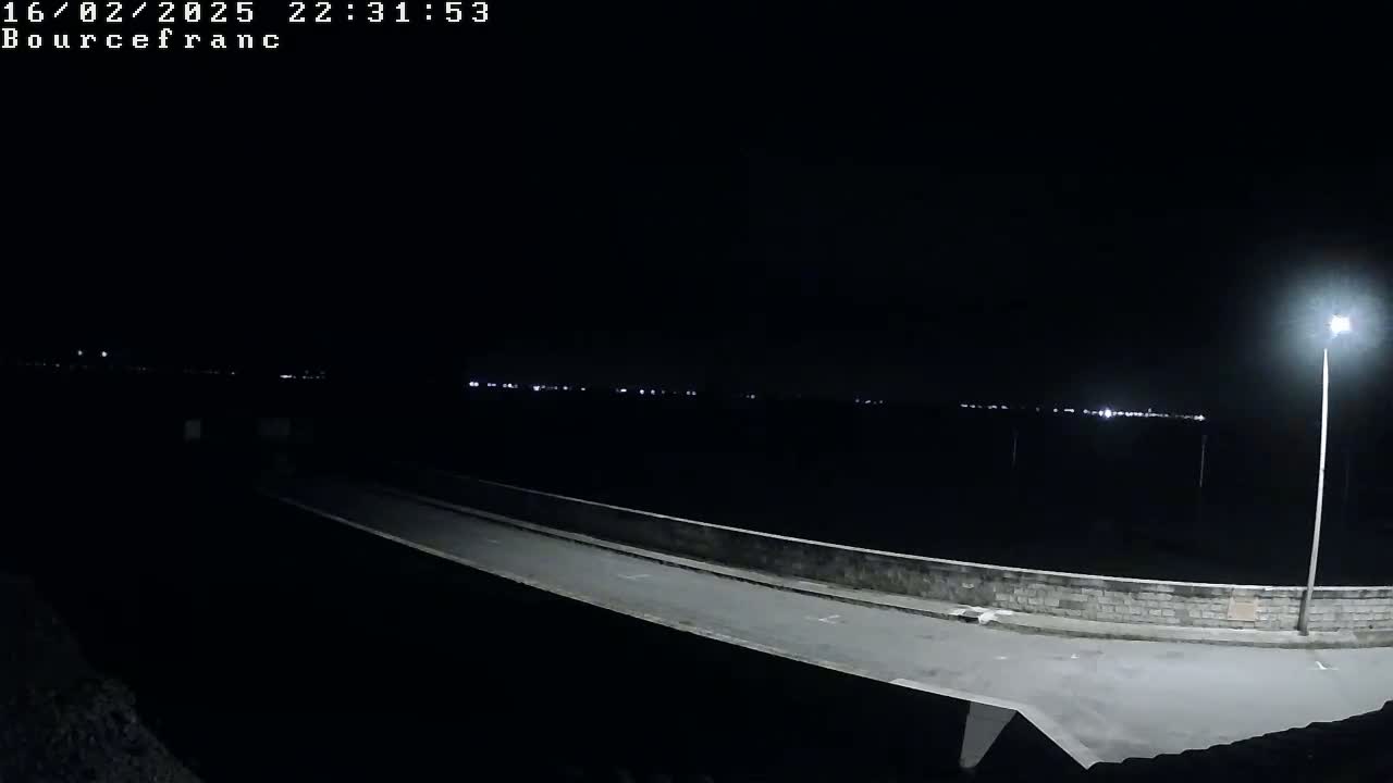 A nighttime view of a paved road leading to a low wall, with distant lights visible in the dark, under a clear sky.