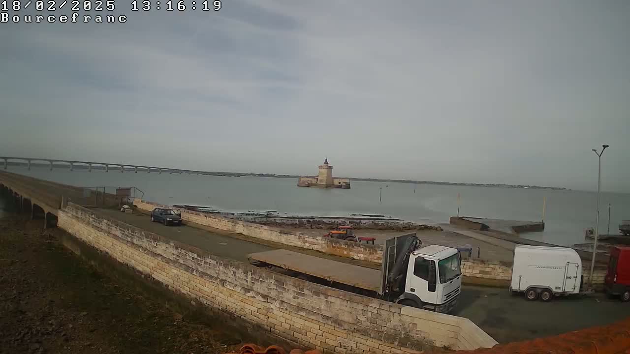 A stone seawall with trucks and a car parked on it overlooks a calm body of water, a distant bridge, and a small stone tower in the middle of the water under an overcast sky.