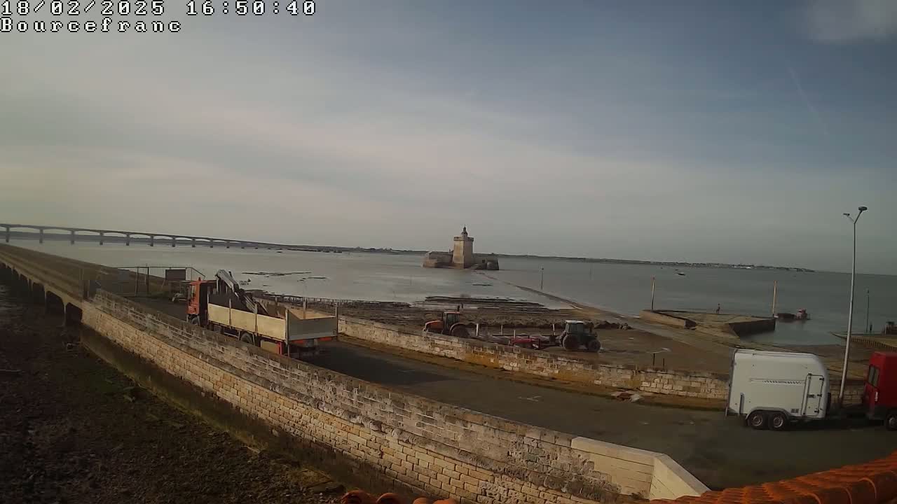 A partly cloudy day shows construction vehicles near a stone seawall and a distant tower, with a bridge visible in the background.