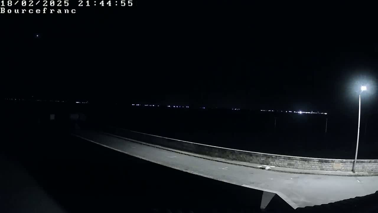 A nighttime view of a paved roadway with a low wall, illuminated by a single lamppost, overlooks a dark expanse where distant lights are visible.