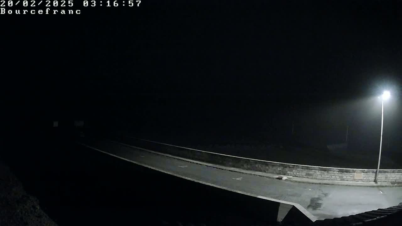 A nighttime, foggy view of a paved road with a low wall on either side, illuminated by a single lamppost.